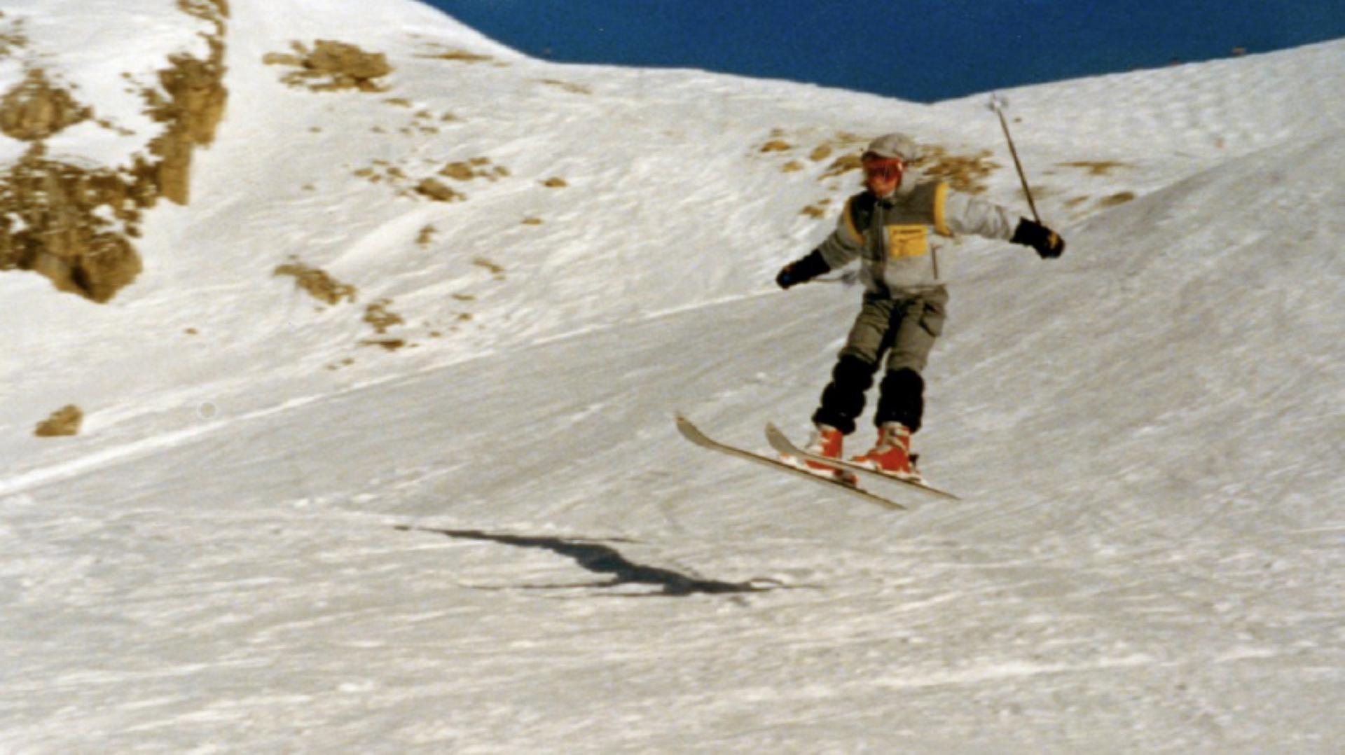 A young Dyson boy on a ski slope, making a jump, with ski poles in the air