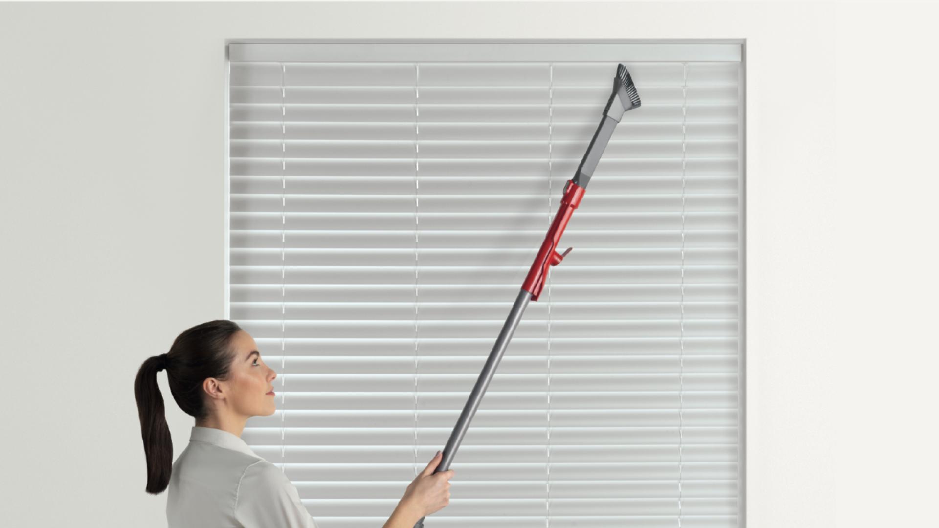 Woman using vacuum wand to clean the top of a blind that's otherwise out of reach.